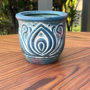 May include: A small, round ceramic planter with a dark blue exterior and a decorative white floral pattern. The rim has a red accent. The planter is sitting on a wooden surface. The interior is green.