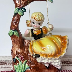 May include: A vintage ceramic figurine of a girl on a swing. The figurine features a girl in a yellow dress with black accents, seated on a swing attached to a brown tree-like structure. The base has green leaves and pink flowers.