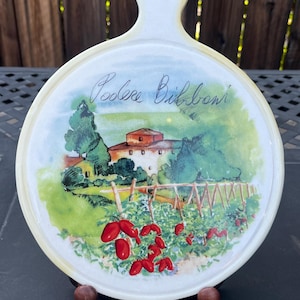 May include: White ceramic cutting board with a handle, featuring a watercolor painting of a countryside scene with a house, trees, and tomato plants. The board has the text "Poolee Bibboni" written on it.