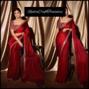 Puede incluir: Un sari rojo vibrante con intrincados adornos plateados en los bordes y el escote. El sari presenta una blusa a juego con bordados detallados. La imagen incluye el texto "RudraCraftTreasures".