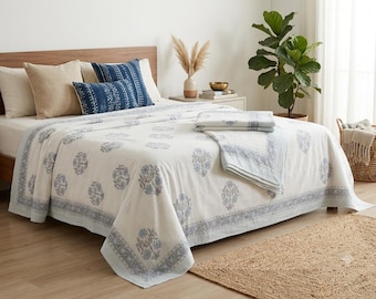 May include: A white bedspread featuring a repeating blue floral pattern and a wide decorative border. The set includes two solid beige square pillows and two rectangular blue pillows with white geometric designs, all arranged on a bed.
