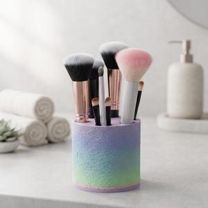 Könnte beinhalten: Ein zylindrischer Make-up-Pinselhalter mit einer texturierten Farbverlauf-Oberfläche, die von Violett oben zu Blau und Grün unten übergeht. Der Halter enthält mehrere Make-up-Pinsel mit unterschiedlichen Borstenfarben, darunter Schwarz, Weiß und Rosa, sowie metallische Griffe in Roségold, Silber und Schwarz.