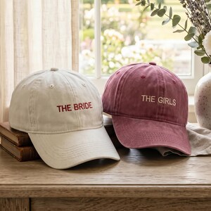 Peut inclure: Deux casquettes de baseball, une blanche et une bordeaux, sont posées sur une surface en bois. La casquette blanche porte l'inscription "THE BRIDE" brodée en rouge, tandis que la casquette bordeaux indique "THE GIRLS". Les deux casquettes sont décontractées et de conception classique.