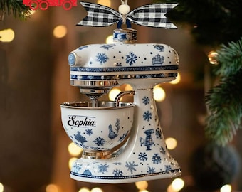 May include: White and blue ceramic stand mixer Christmas ornament. The ornament features a bowl with the name "Sophia" and is decorated with blue snowflakes and other festive designs. A black and white plaid bow is at the top.