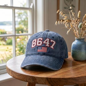 Könnte beinhalten: Eine blaue Baseballkappe aus Denim mit der Zahl "8647" in Weiß und Rot gestickt, zusammen mit einer kleinen amerikanischen Flagge. Die Kappe liegt auf einem Holztisch in der Nähe eines Fensters.