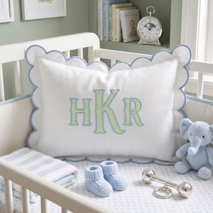 May include: A rectangular white pillow with a scalloped edge trimmed in light blue piping. The center features an embroidered monogram with the letters HKR in light green with a dark blue outline. The pillow is placed inside a white crib.