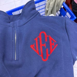 May include: Navy blue quarter-zip sweatshirt with a red embroidered monogram. The sweatshirt features a high collar and a silver zipper. The red monogram is in a diamond shape.