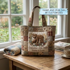 Puede incluir: Un bolso tote de patchwork con un diseño de familia de osos. La bolsa presenta un panel central bordado con una osa y dos cachorros, rodeados de detalles florales. El texto dice "Grandma - Est. 1995 Home is Love". La bolsa tiene un diseño a cuadros y patchwork.