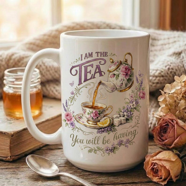 May include: White ceramic mug with a large handle, featuring a floral design and the text "I AM THE TEA. You will be having." The design includes a teapot, teacup, and floral accents. A spoon, jar, and roses are in the background.