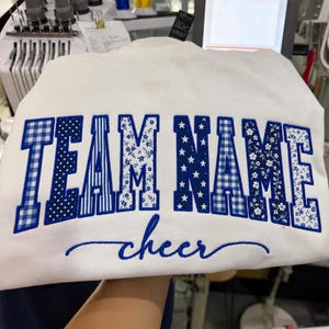 Könnte beinhalten: Ein weißes Sweatshirt mit dem gestickten Text "TEAM NAME" in einer fetten Blockschrift. Jeder Buchstabe ist mit einem anderen blau-weißen Muster gefüllt, darunter Vichy-Karo, Punkte, Streifen, Sterne und Blumen. Das Wort "cheer" ist darunter in blauer Schreibschrift gestickt.