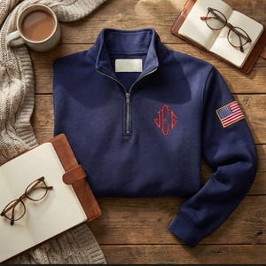 May include: Navy blue quarter-zip sweatshirt with a red embroidered monogram and an American flag patch on the sleeve. The sweatshirt has a blank white label on the collar and a silver zipper. The background is a wooden surface.