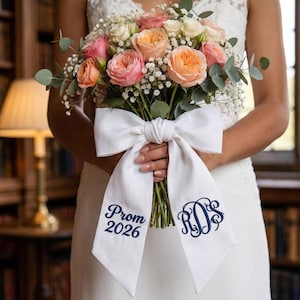 May include: A bridal bouquet featuring peach and pink roses, white baby's breath, and greenery. The bouquet is tied with a large white bow with the text "Prom 2026" and a monogram in navy blue.