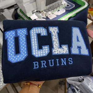 Könnte beinhalten: Ein marineblaues Sweatshirt mit dem Schriftzug UCLA in großen, applizierten Buchstaben mit verschiedenen hellblauen Mustern wie Punkten, Vichy-Karos, Blumen und Streifen. Unter den großen Buchstaben ist das Wort BRUINS hellblau gestickt.