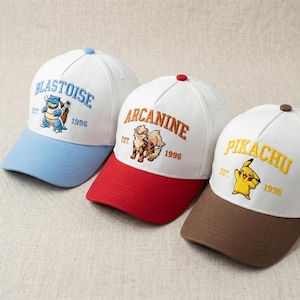 May include: Three white baseball caps with embroidered Pokémon character designs. Each cap features a different character: Blastoise, Arcanine, and Pikachu. The caps have coloured brims in blue, red, and brown, with the year 1996.