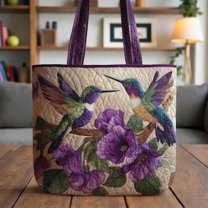 May include: A quilted tote bag featuring a detailed design of two hummingbirds with vibrant purple, green, and blue plumage, perched near purple flowers. The bag has a cream-colored background, a quilted pattern, and purple handles.