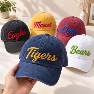 Op de afbeelding: Een collectie van vijf baseballpetten in zwart, geel, bordeaux, marineblauw en wit. Elke pet is voorzien van geborduurde tekst op de voorzijde: Eagles in rood, Mascot in roze, Lions in blauw, Tigers in geel en Bears in groene letters.