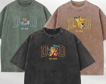 May include: Three mineral-washed t-shirts in sage green, brown, and charcoal gray. Each features a large letter D on both sides of a Pokémon graphic with text below: EST. 2026, EST. 2016, and EST. 23/25 respectively. The shirts have crew necks.