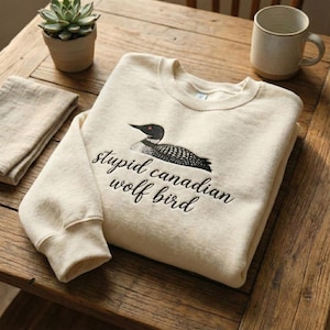 May include: Cream-colored sweatshirt featuring an embroidered loon design with the text "stupid canadian wolf bird" in black. The sweatshirt is folded on a wooden surface, with a small plant and mug in the background.