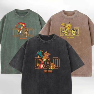 May include: Three mineral-washed crewneck t-shirts in green, brown, and black. Each features the word DAD in large orange or yellow letters, accompanied by various Pokémon characters and an establishment year printed below the text.