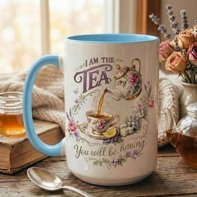 May include: White ceramic mug with a light blue handle and rim, featuring a floral tea-themed design. The mug has the text "I AM THE TEA" and "You will be having". A teapot pours tea into a teacup, surrounded by flowers.
