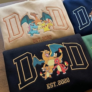 May include: A beige sweatshirt with embroidered DAD text, Charizard, Totodile, and Pikachu, featuring EST. 2022. A black sweatshirt with embroidered DAD text, Charizard, Charmander, and Charmelon, featuring EST. 2020. A navy sweatshirt with a partial D.
