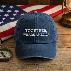 May include: A navy blue baseball cap with the text "TOGETHER, WE ARE AMERICA" in white. The cap is set against a backdrop of an American flag, a compass, and a vintage lamp on a wooden surface.
