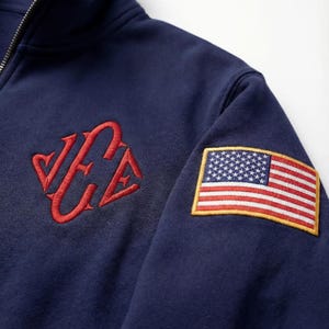 Könnte beinhalten: Marineblaues Sweatshirt mit Viertelreißverschluss, rotem gesticktem Logo und einem amerikanischen Flaggenaufnäher auf dem Ärmel. Das Sweatshirt hat einen silbernen Reißverschluss.