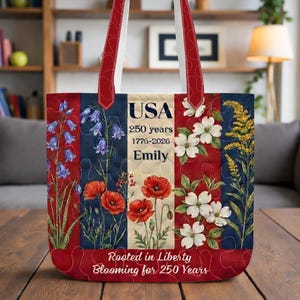 Pode incluir: Uma sacola patriótica com um design floral vermelho, branco e azul e texto. A sacola apresenta a palavra "USA", o texto "250 anos 1776-2026 Emily" e a frase "Rooted in Liberty Blooming for 250 Years".
