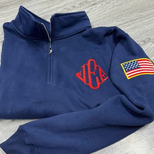 May include: Navy blue quarter-zip sweatshirt with a red monogram on the chest and an American flag patch on the sleeve. The sweatshirt has a high collar and a silver zipper.