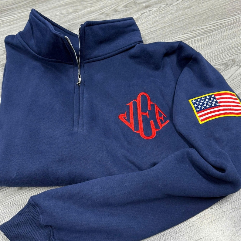 May include: Navy blue quarter-zip sweatshirt with a red monogram on the chest and an American flag patch on the sleeve. The sweatshirt has a high collar and a silver zipper.