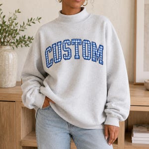 May include: A light grey mock neck sweatshirt featuring the word CUSTOM in a blue and white gingham check pattern across the chest. The garment has long sleeves with ribbed cuffs and a ribbed hem, constructed from a soft, heathered fabric.