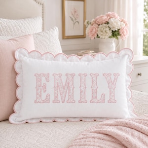 May include: A rectangular white throw pillow with a scalloped edge trimmed in light pink. The center of the pillow features the name EMILY embroidered in large, light pink capital letters with a delicate floral pattern inside the lettering.