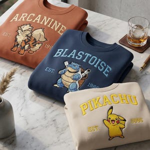 Peut inclure: Trois pulls à col rond en rouille, bleu marine et crème. Chacun présente un personnage Pokémon brodé, le nom du personnage en lettres arquées et le texte EST. 1996. Le pull rouille montre Arcanin, le bleu marine Tortank et le crème Pikachu.