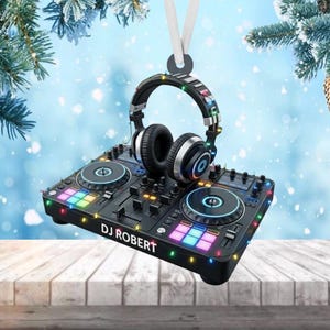 May include: A black DJ controller Christmas ornament with a pair of headphones. The ornament has colorful lights around the edges and the text "DJ ROBERT". The background is a winter scene with falling snow.