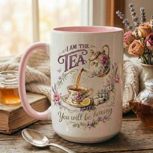 May include: White ceramic mug with a pink handle and interior, featuring a floral tea-themed design. The mug has the text "I AM THE TEA" and "You will be having". A spoon, book, and jar of honey are in the background.