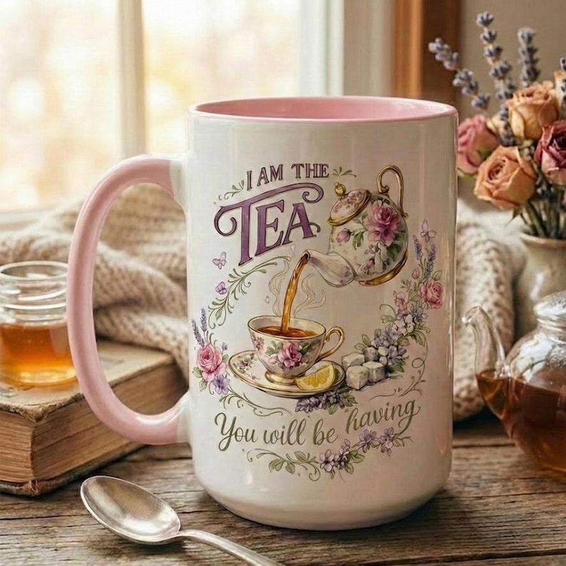 May include: White ceramic mug with a pink handle and interior, featuring a floral tea-themed design. The mug has the text "I AM THE TEA" and "You will be having". A spoon, book, and jar of honey are in the background.