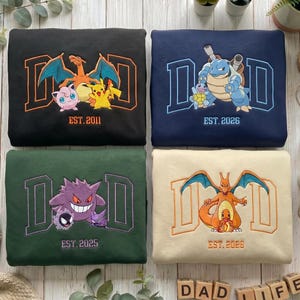 May include: Four folded sweatshirts featuring embroidered designs. The black sweatshirt shows Charizard, Pikachu, and Jigglypuff with 'DAD EST. 2011'. The navy sweatshirt shows Blastoise and Squirtle with 'DAD EST. 2026'. The green sweatshirt shows Gengar, Gastly, and Haunter with 'DAD EST. 2025'. The cream sweatshirt shows Charizard and Charmander with 'DAD EST. 2026'.