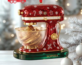 May include: A red and gold Christmas ornament shaped like a stand mixer. The ornament has snowflake, candy cane, and gingerbread man designs. The base is green with the name "SOPHIA" and the year "2025".