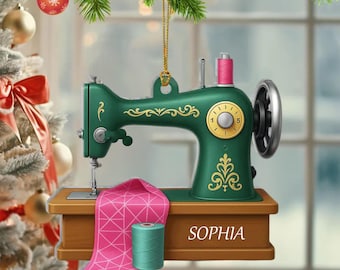 May include: A green sewing machine Christmas ornament with gold accents, a pink spool of thread, and a pink fabric. The ornament has the name "SOPHIA" on the base. Festive holiday decor.