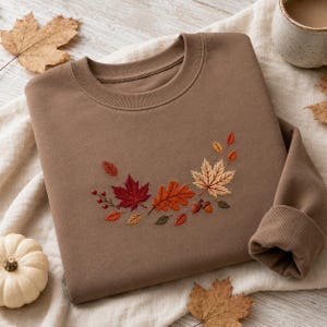 Op de afbeelding: Een bruine sweater met ronde hals voorzien van een geborduurd ontwerp van herfstbladeren, waaronder rode esdoornbladeren, oranje eikenbladeren en kleine bruine eikeltjes in een gebogen patroon in het midden van de borst.