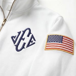 May include: White zip-up sweatshirt with a navy blue embroidered logo and an American flag patch on the sleeve. The sweatshirt features a gold zipper and a clean, minimalist design.