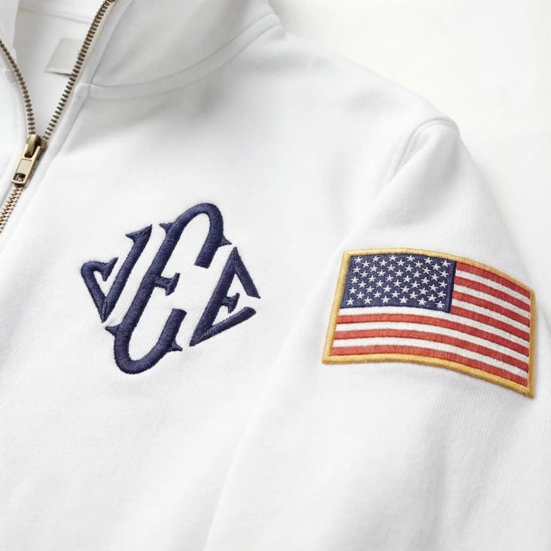 May include: White zip-up sweatshirt with a navy blue embroidered logo and an American flag patch on the sleeve. The sweatshirt features a gold zipper and a clean, minimalist design.