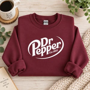 May include: A maroon long-sleeve crewneck sweatshirt featuring the white Dr Pepper logo with the text Est.1885 printed in the center. The garment has a Gildan brand label on the inside collar and is made of a soft, fleece-lined fabric material.