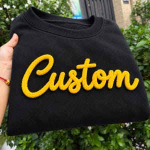 Puede incluir: Una sudadera negra de cuello redondo con la palabra Custom en una fuente cursiva de color amarillo brillante, texturizada y en relieve en el pecho. Las letras tienen un aspecto grueso similar al hilo que destaca sobre la tela oscura.