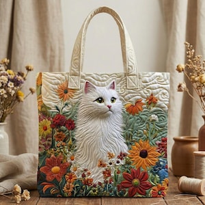 May include: Cream-colored tote bag featuring a detailed, quilted design of a white cat amidst colorful flowers. The bag has a quilted handle and a textured background. The flowers include orange, red, and yellow blooms, creating a vibrant, artistic aesthetic.