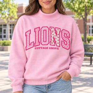Puede incluir: Una sudadera de manga larga en color rosa claro con cuello alto. El frente presenta la palabra LIONS en letras grandes con variados patrones rosas y blancos, incluyendo cuadros vichy, rayas y estampados florales, con COTTAGE GROVE debajo.
