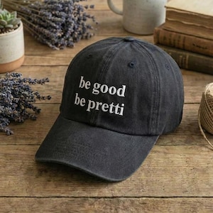May include: A dark grey baseball cap with the embroidered text "be good be pretti" in white. The cap has a curved peak and a classic six-panel design. The hat is made of a soft, durable fabric.