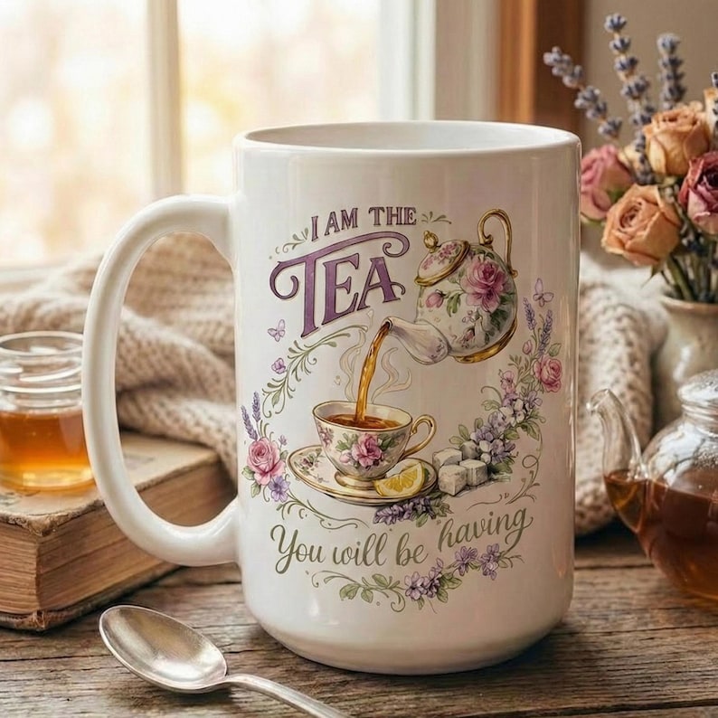 May include: White ceramic mug with a large handle, featuring a floral design and the text "I AM THE TEA. You will be having." A teapot pours tea into a teacup, surrounded by flowers and a lemon slice. The mug is on a wooden surface.