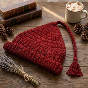 Op de afbeelding: Een handgemaakte, rode gehaakte muts met een lange, gevlochten kwast. De muts is gemaakt van een getextureerd garen en heeft een puntige vorm. De kwast eindigt in een decoratieve franje. De muts wordt gepresenteerd op een houten oppervlak.