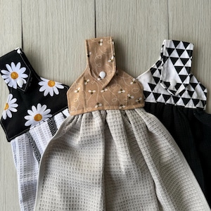May include: Three kitchen towels with different fabric patterns. The towels have a loop at the top with a button closure. The patterns are black and white daisies, brown with bees, and black and white triangles.
